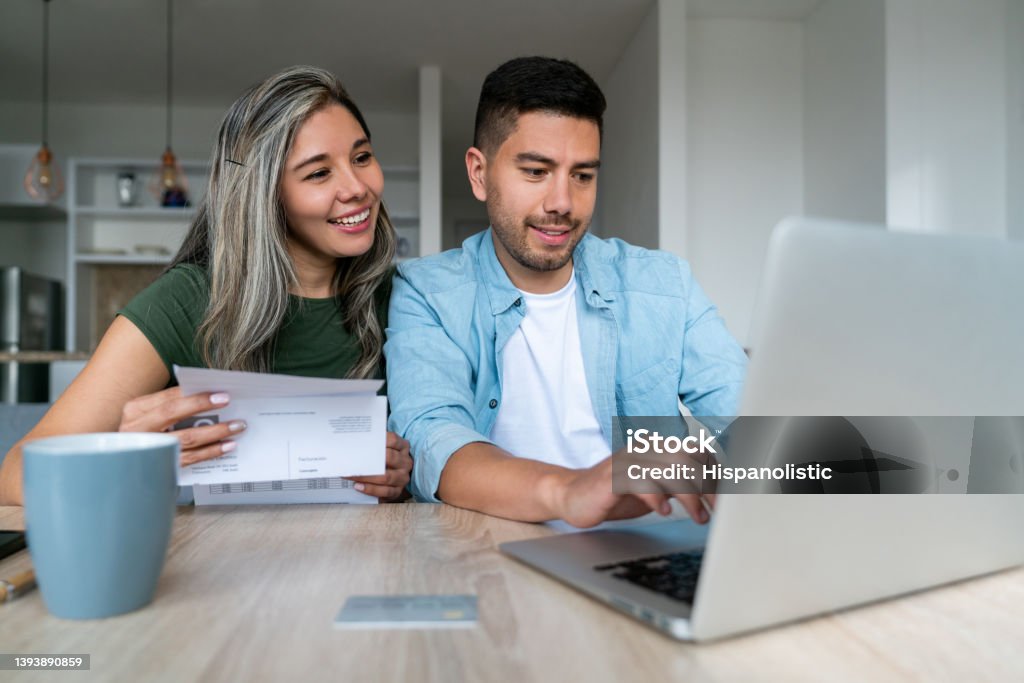 Happy couple at home paying bills online Happy Latin American couple at home paying bills online on their laptop and smiling - financial technology concepts Financial Bill Stock Photo