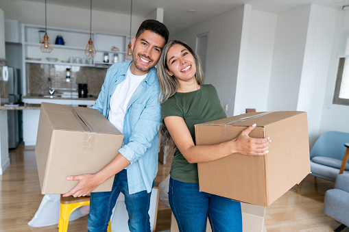 Portrait of a happy Latin American couple packing in boxes while moving house and looking at the camera smiling