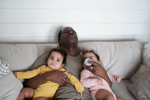 Tired Parent Falling Asleep High angle view of exhausted mature African American dad falling asleep while taking care of his baby twin daughters sleeping grandpa stock pictures, royalty-free photos & images