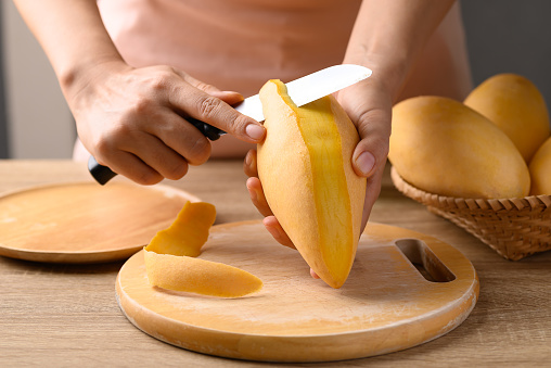 Hand holding knife and peeling Thai mango fruit (Nam Dok Mai) on wooden board, Tropical fruit in summer season
