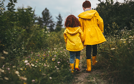 Back view of unrecognizable redhead brother and sister wearing vivid yellow rubber boots and waterproof raincoats walking along path in meadow