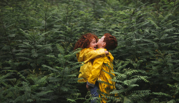 Side view of smiling redhead bother lifting little sister in yellow raincoat while standing in green forest