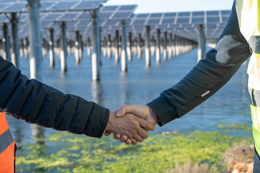 Two male workers shake hands in front of the seaside solar power station