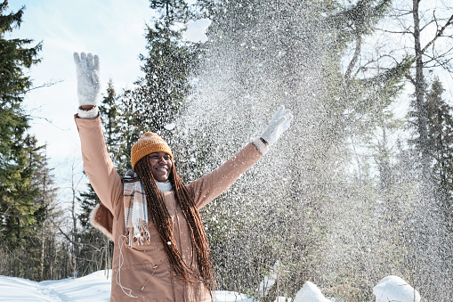Playful young African American woman with long hair having fun throwing snow in the air on sunny winter day