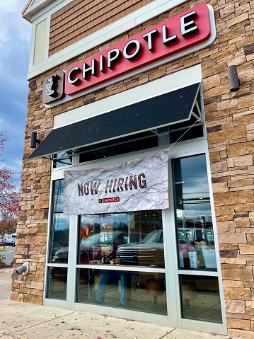 Fairfax, Virginia, USA - November 29, 2021: A “Now Hiring” banner hangs from the awning of a Chipotle fast casual restaurant in the City of Fairfax as a man eats his meal inside.