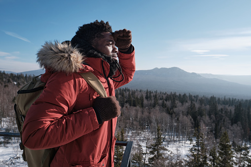 Young African American man wearing backpack standing at viewing platform on mountain top on winter day looking into distance