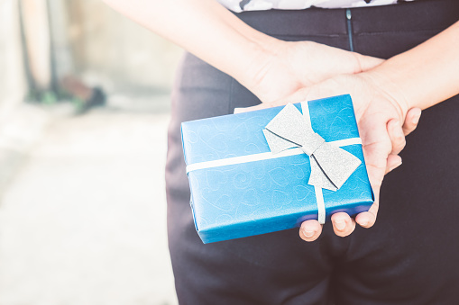 A woman hiding package presents for her boyfriend behind the back. Surprising blue gift box wrapped in ribbon bow. Merry Christmas, Happy new year or Anniversary birthday, Valentine's day. Copy space