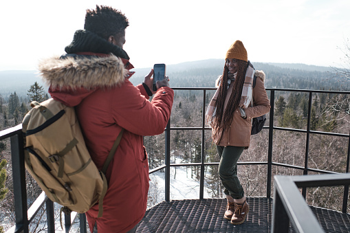 Young Black male tourist standing at observation deck on mountain top taking photos of his beautiful girlfriend