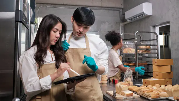 Three young friends and startup partners of bread dough and pastry foods busy with homemade baking jobs while cooking orders online, packing, and delivering on bakery shop, small business entrepreneur.