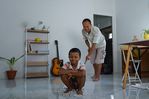 Father and son cleaning in living room at home.
A son sit on the mop and laughed. while father was mopping the floor.