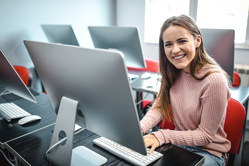 Happy female student using desktop PC in an empty computer lab and looking at camera.