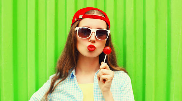 Portrait of young woman with lollipop blowing her lips sends kiss wearing red baseball cap on green background stock photo