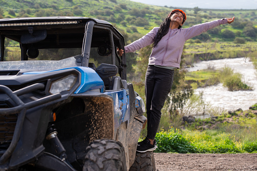 Happy woman with raised hand standing on wheel of the off-road SxS vehicle, enjoying the scenic view of mountain river after a successful ride. The woman wearing UTV Powersports goggles. An extreme, adrenaline-pumping adventure on the terrain, riding tour on a 4x4, Side-by-Side, UTV vehicle. Outdoor recreation activities. Off-Road Experience.