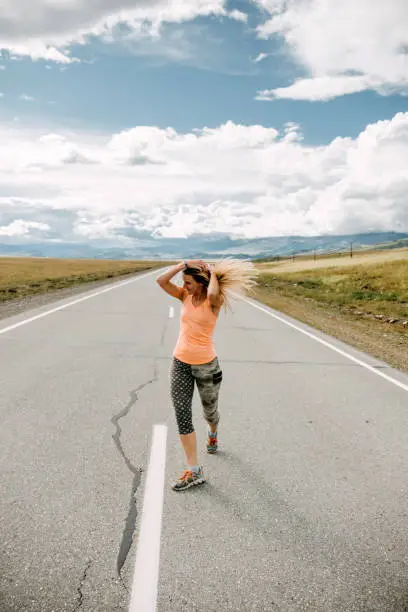 A young blonde woman in sportswear stands in the middle of a car-free road among the mountains and straightens her hair. Travel content