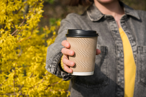Young woman holding kraft paper coffee cup against yellow blooming trees as background. Spring or summer coffee take away concept