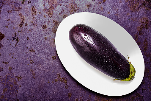 High angle view of delicious raw eggplant washed with fresh clean water on white plate over purple color stone table.