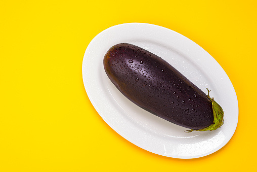 High angle view of delicious raw eggplant washed with fresh clean water on white plate over yellow color background. Image made in studio