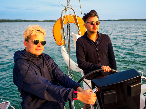 gay couple on a sailboat, one person steering wheel sailing yacht other is standing near bow and looking forward at sunset, front view