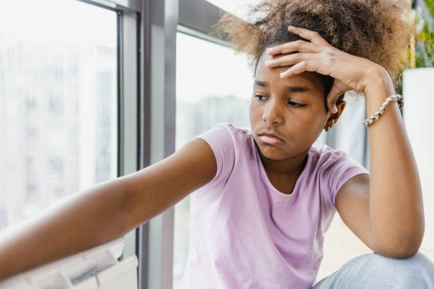 pensativa e triste garota afro-americana sentada em casa perto da janela - só meninas adolescentes - fotografias e filmes do acervo