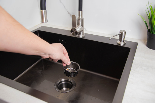 A man's hand removes a metal strainer from a kitchen sink drain. Cleaning the drain and pipes from clogging with food particles after washing dishes, close-up