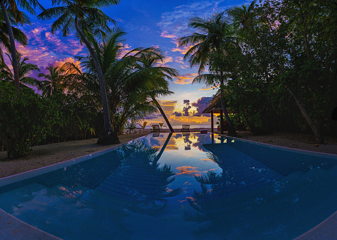 Luxury sunset over infinity pool in a summer beachfront hotel resort at tropical landscape. Tranquil beach holiday vacation background mood. Amazing island sunset beach view, palms swimming pool