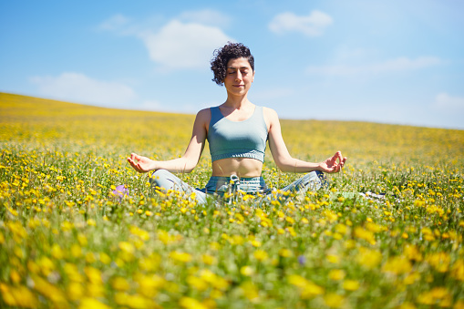 Young woman meditating and practising yoga in a meadow full with colorful wildflowers and blue sky in a sunny day. Soul-searching and spiritual relaxation in nature