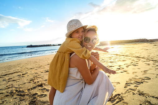 Happy mother and her little daughter having fun together on the sandy beach during sunset. Lovely kid embracing and playing with her mom during summer vacation. Real people emotion. Family lifestyle.