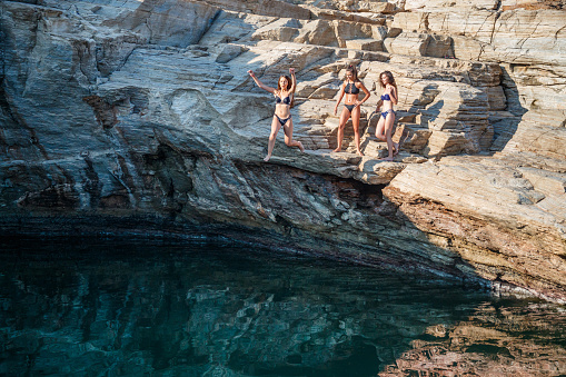 Young woman in bikini diving on her feet into sea water in natural pool from rocky cliff, her friends watching her from rock