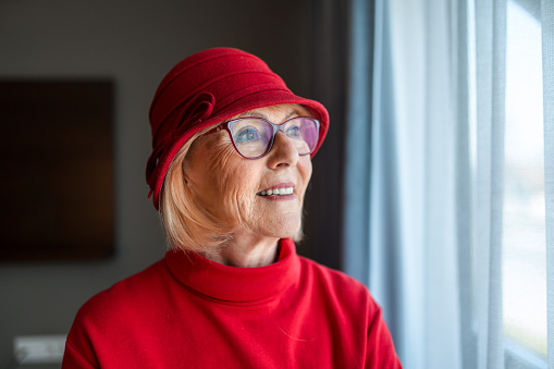 An older woman smiles as she stares out of her hotel room window. Natural light is hitting her face through sheer curtains. She is dressed in a red dress, hat, and glasses.