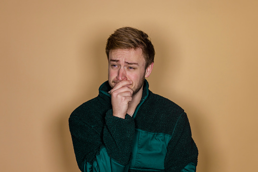 A front view waist-up portrait of a young caucasian man expressing negative emotion and concern, he has his hand on his mouth and he is crying, he is in a studio standing in front of a cream background.