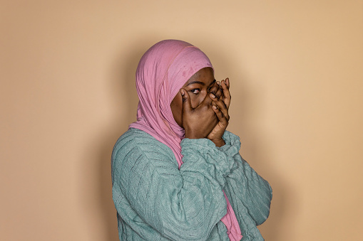 A front view waist up portrait of a young black woman standing in front of a cream background with her hands covering her mouth, she is looking at the camera with a shocked emotion.