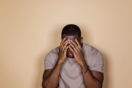 A front view waist-up portrait of a young black man expressing negative emotion and concern, he has his hands over his face, he is in a studio in front of a cream background.