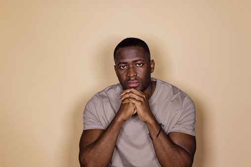 A front view waist-up portrait of a young black man expressing negative emotion and concern, he has his hand on his chin, he is in a studio in front of a cream background.
