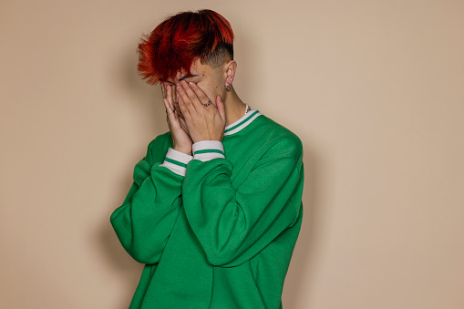 A front view waist-up portrait of a young Asian man expressing negative emotion and concern, he has his hands over his face, he is in a studio in front of a cream background.