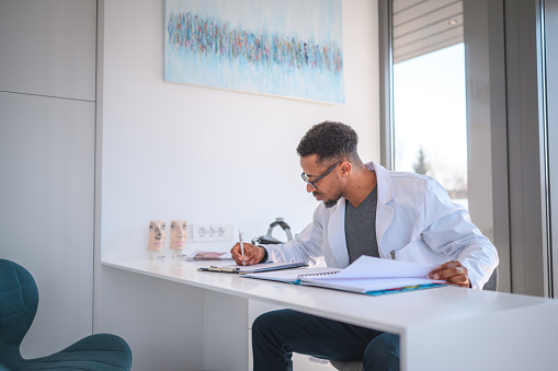 African American doctor siting in a bright modern office in a new clinic. Making notes and writing them in a medical chart.