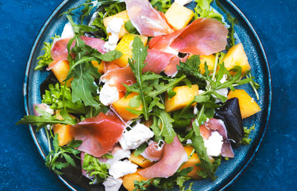 Delicious summer fresh salad with cantaloupe melon, prosciutto, soft cheese and arugula on blue table background, top view, copy space stock photo