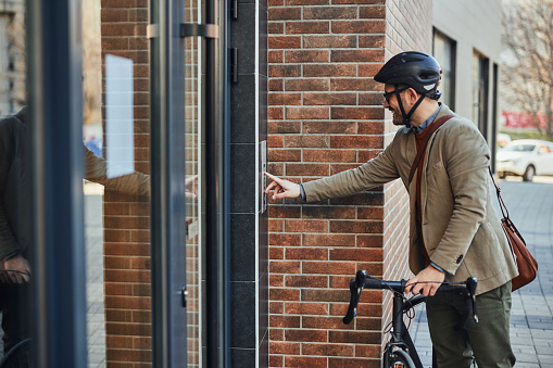 Businessman cyclist with helmet standing in front of office building and rings on intercom