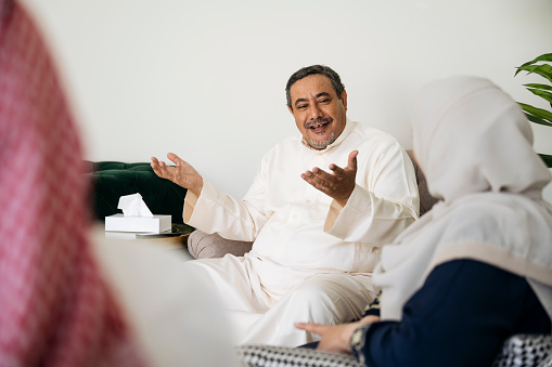 Over the shoulder view of man in dish dash smiling and gesturing as he converses with woman in abaya and headscarf.