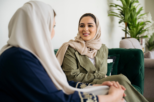 Side view of late 20s and late 40s women, each wearing traditional Saudi abaya and hijab, smiling face to face as they enjoy weekend leisure time.