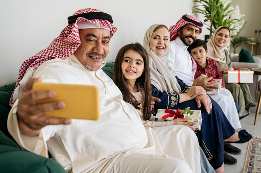 Side view of three-generation Saudi family in traditional attire and western dress sitting on sofa and smiling as mature man captures memory with smart phone.