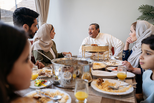 Over the shoulder view with focus on Middle Eastern adults smiling and talking as they enjoy traditional dishes at dining table in modern Riyadh home.