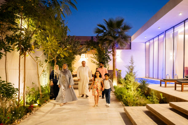 Three-generation Middle Eastern family outside family home Full length view of approaching men, women, and children in traditional and western dress smiling at camera outside illuminated modern house at twilight. blue hour twilight stock pictures, royalty-free photos & images