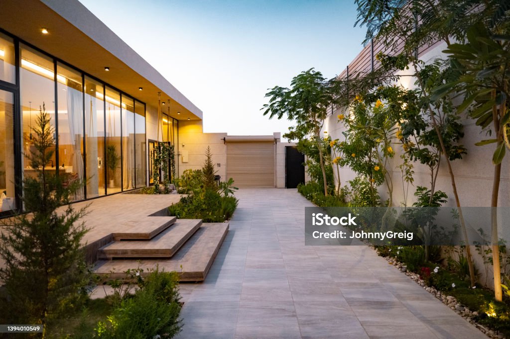Home exterior at dusk in Riyadh, Saudi Arabia Diminishing perspective outside illuminated modern home with full length glass windows, steps to entrance, and native plant landscaping. Landscaped Stock Photo