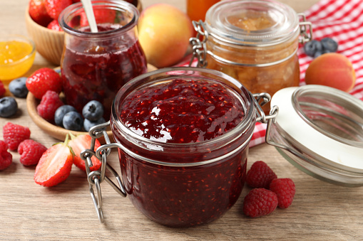 Jars with different jams and fresh fruits on wooden table, closeup