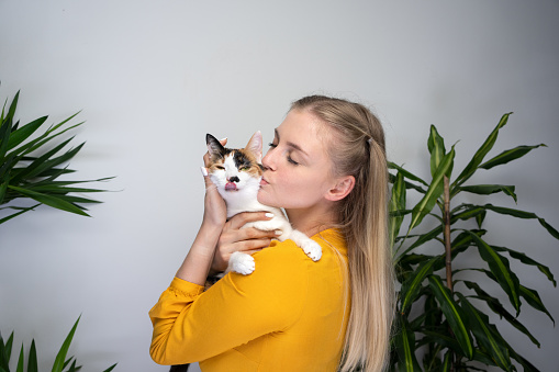 young blond woman carrying her cat kissing it on the head. the cat dislikes it and is sticking out tongue.