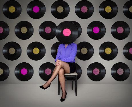 Conceptual photomanipulation of woman sitting on a chair with a record over her face, against the wall full of records. 
The illustration of: The irony of woman's nature, relation difficulties, communication problems, rigidity, no will of change, telling the same stories over and over, same story syndrome.