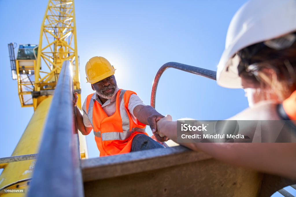 African american male cargo handler helping female coworker to getting on harbor cargo crane Low angle view of african american male cargo handler helping female coworker to getting on harbor cargo crane during daytime Ladder Stock Photo