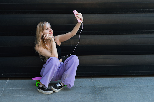 A blond teenage girl with street-style clothing is chilling on her skateboard in front of a black wall. She is listening to music on her phone and taking a selfie.