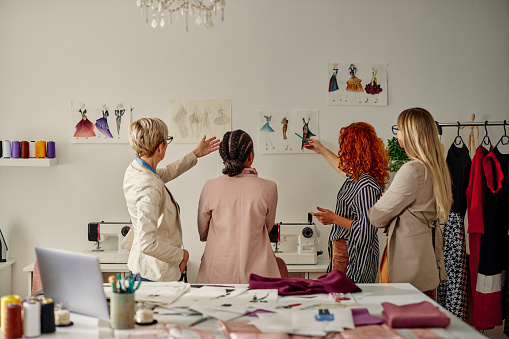 View from behind of diverse multi-generations fashion designers pointing on sketches which hanging on the wall and talking about project plans.