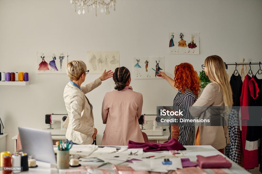 Four female coworkers working on their fashion design View from behind of diverse multi-generations fashion designers pointing on sketches which hanging on the wall and talking about project plans. Fashion Stock Photo